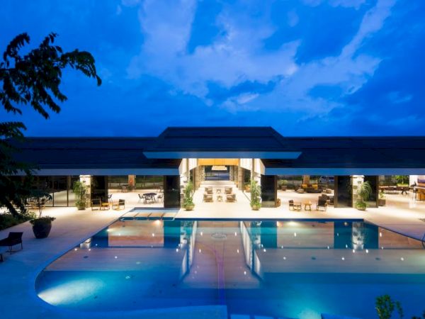 A modern building with large windows is lit up at night, overlooking a large rectangular swimming pool reflecting the blue evening sky.