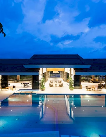 A luxurious building with a large illuminated swimming pool in front, under a blue evening sky, surrounded by trees.