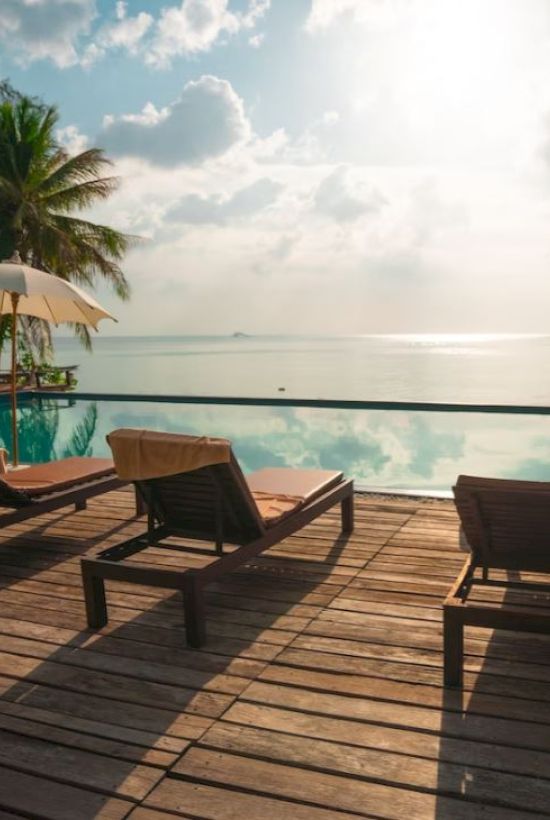 The image shows a serene seaside view with lounge chairs, umbrellas, and a pool, overlooking a calm ocean under a partly cloudy sky.