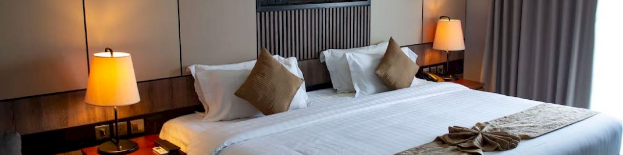 The image shows a neatly made hotel room with a large bed, lamps, and curtains, creating a cozy and inviting atmosphere.