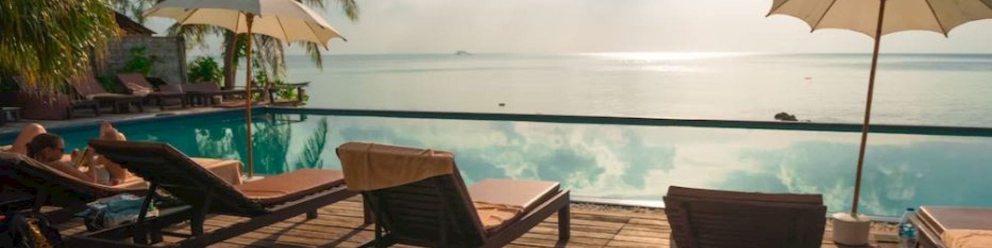 The image shows a serene poolside scene with lounge chairs, umbrellas, and a scenic view of the ocean under a partly cloudy sky.