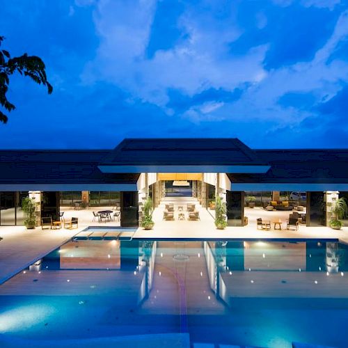The image shows a modern building with a swimming pool in the foreground, surrounded by lounge chairs, under a blue, evening sky.