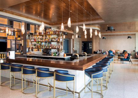 The image shows a modern bar with high-backed blue stools, a variety of bottles on shelves, and hanging pendant lights.