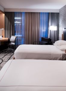 The image shows a modern hotel room with two beds, a TV, a desk, and soft lighting, creating a cozy and inviting atmosphere.