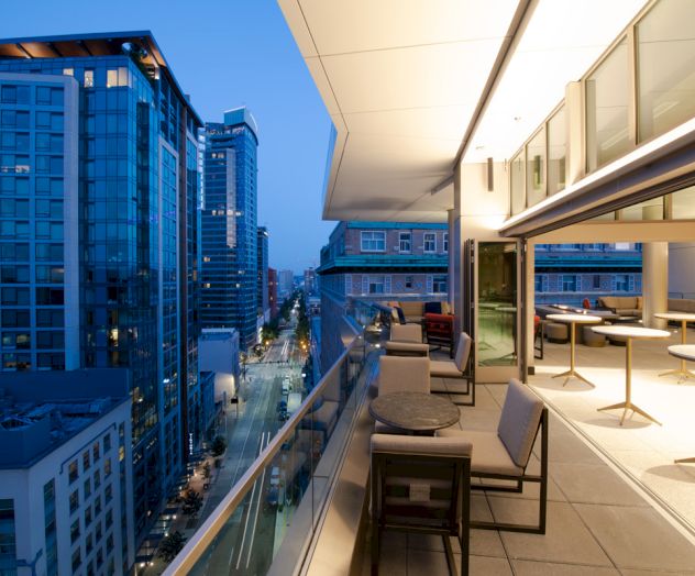 A modern rooftop terrace with seating overlooks a cityscape at dawn or dusk, featuring high-rise buildings and a lit street below.