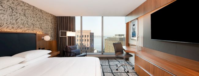The image shows a modern hotel room with a large bed, a wall-mounted TV, a desk, and a large window offering a city view.