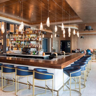 The image shows a modern bar with blue stools, a wooden counter, hanging lights, shelves with bottles, and seating areas in the background.