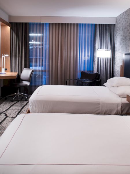 A modern hotel room features two neatly made beds, a wall-mounted TV, desk, and stylish decor, under soft lighting from lamps above the headboard.