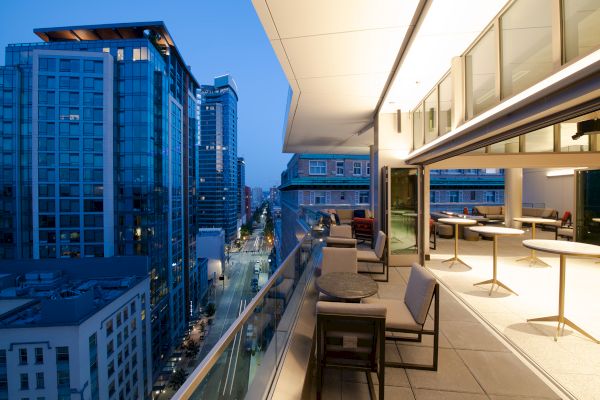A modern urban balcony view featuring patio furniture overlooks a cityscape with tall buildings as evening light gives a serene ambiance.