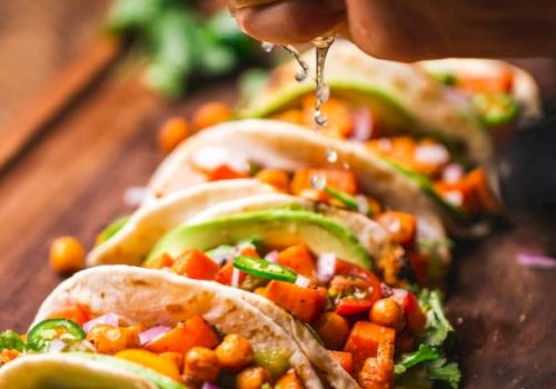A hand squeezes lime over tacos with chickpeas, vegetables, and avocado slices on a wooden board, garnished with lime and cilantro.