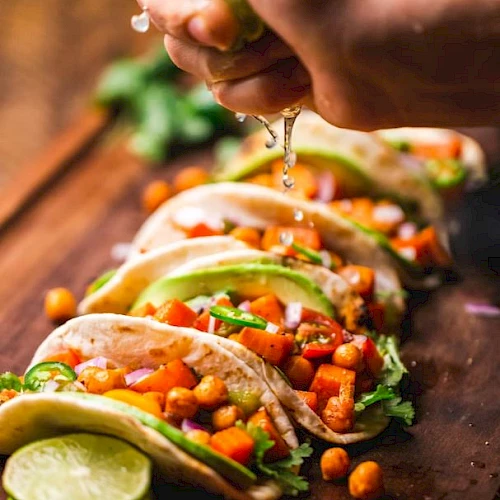 A hand squeezes lime over tacos with chickpeas, vegetables, and avocado slices on a wooden board, garnished with lime and cilantro.