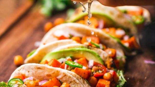 A hand squeezes lime over tacos with chickpeas, vegetables, and avocado slices on a wooden board, garnished with lime and cilantro.