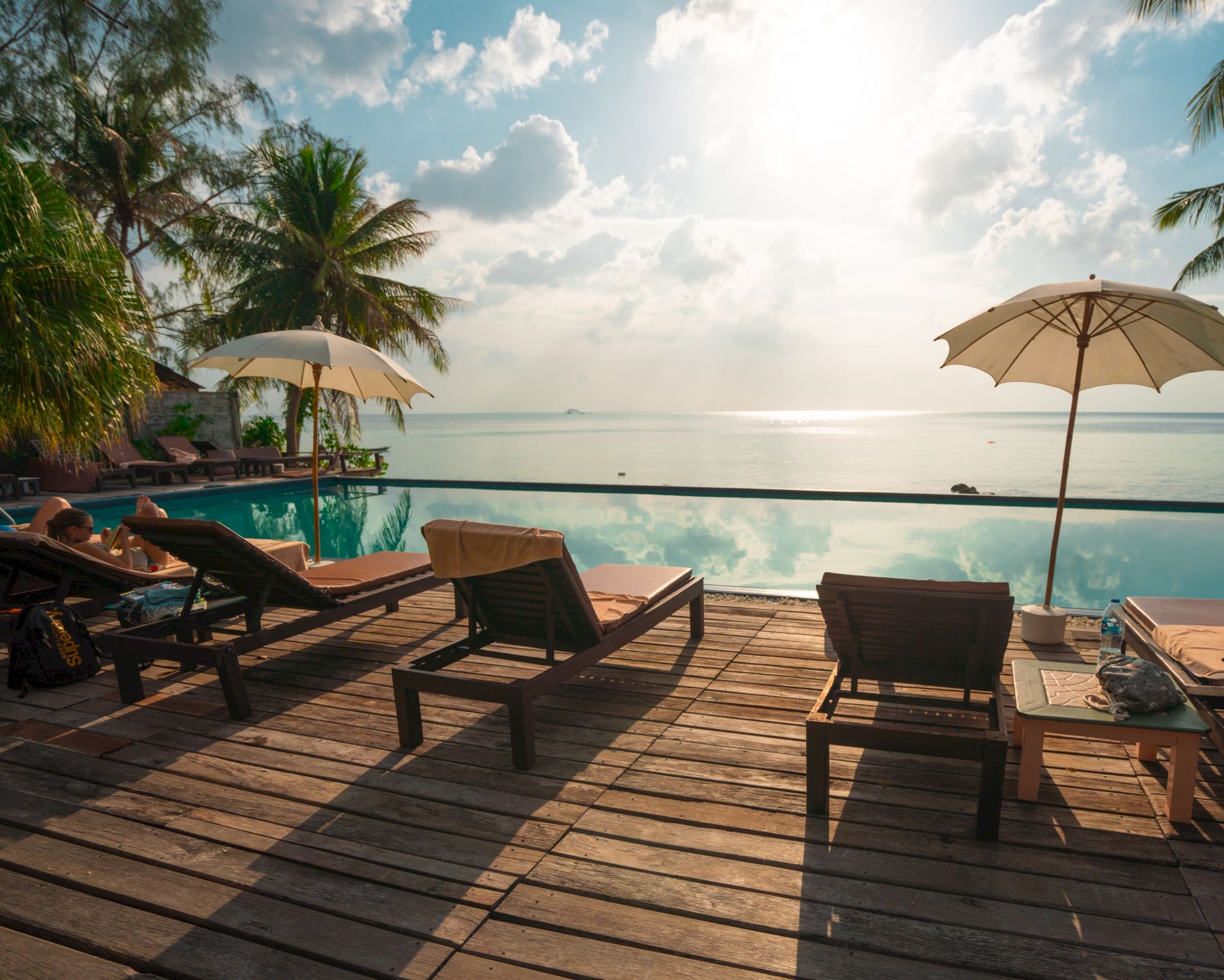 The image shows a peaceful poolside scene with lounge chairs, umbrellas, and a beautiful view of the ocean under a partly cloudy sky.