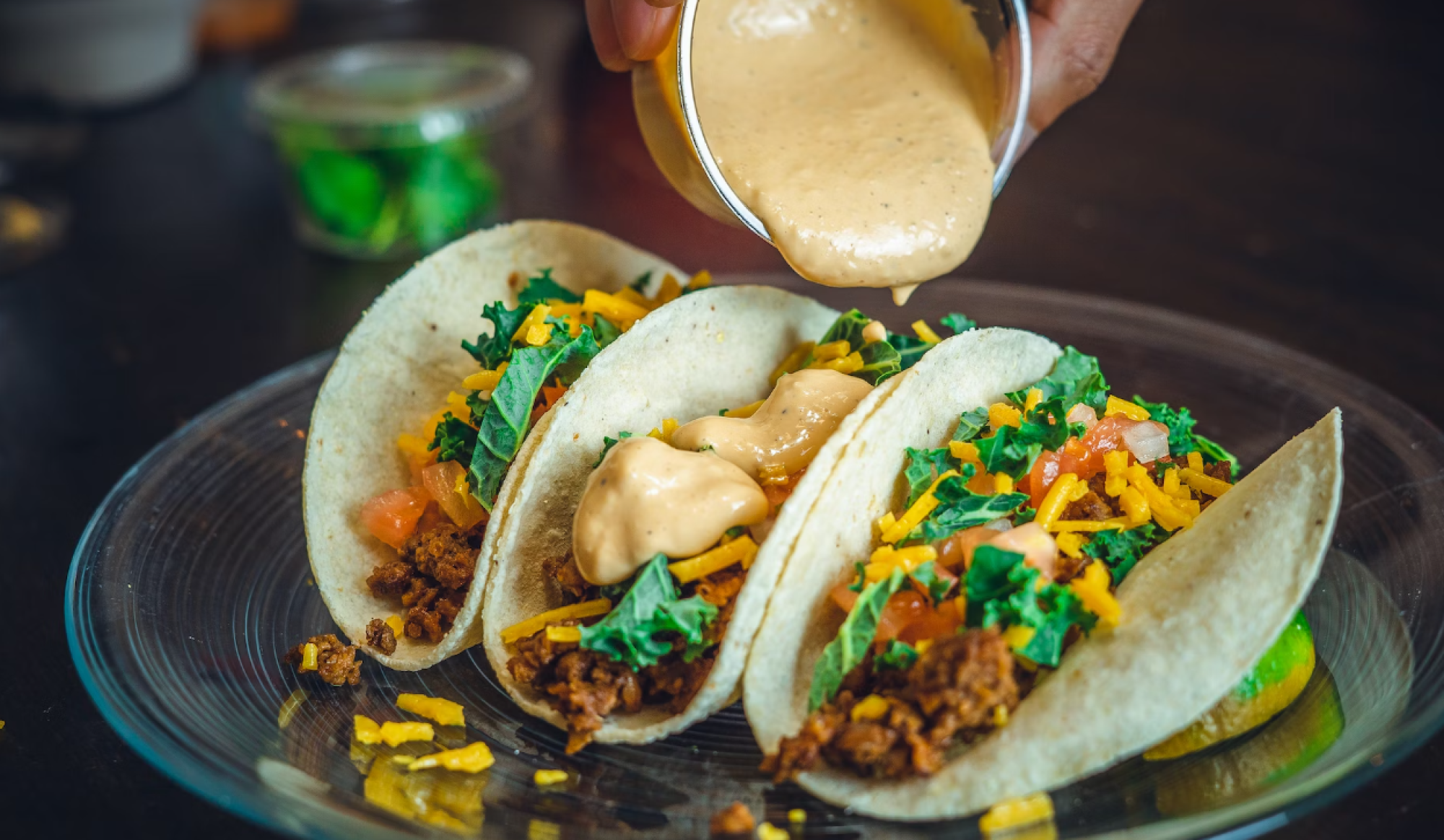 Three tacos on a plate being topped with creamy sauce, filled with meat, shredded cheese, tomatoes, and greens.