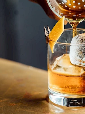 A cocktail being poured through a strainer into a glass with ice and a citrus twist garnish.