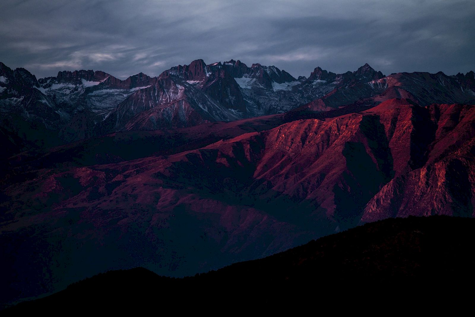 A mountain range is bathed in red light at twilight, with a dramatic sky overhead.