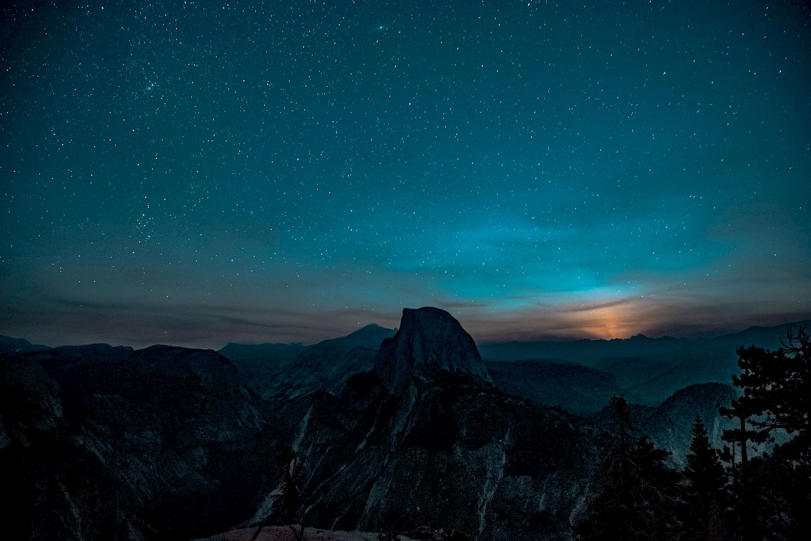 A nighttime landscape shows a starry sky over silhouetted mountains, with a bright glow on the horizon creating a serene scene.