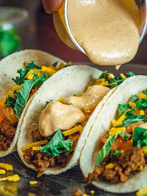 Three tacos filled with ground meat, lettuce, cheese, and sauce being poured on top, displayed on a plate.