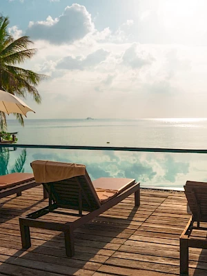 A serene tropical setting with lounge chairs, umbrellas, and a pool, overlooking a calm sea under a partly cloudy sky.
