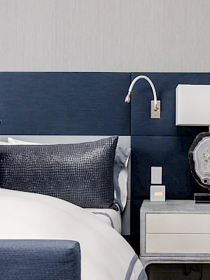 A modern bedroom with a blue headboard, white bedding, nightstand, lamp, and decorative pillows.