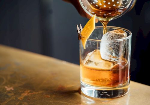 A bartender pours a brown cocktail into a glass with ice and an orange twist garnish, using a strainer.