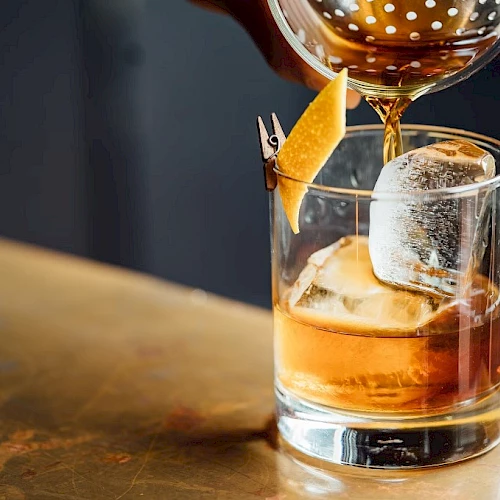 A bartender pours a brown cocktail into a glass with ice and an orange twist garnish, using a strainer.