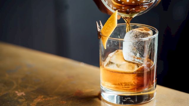 A bartender pours a brown cocktail into a glass with ice and an orange twist garnish, using a strainer.