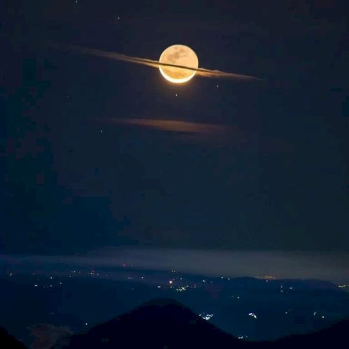 A full moon with clouds appears like Saturn's rings, above a dark landscape with scattered city lights below.