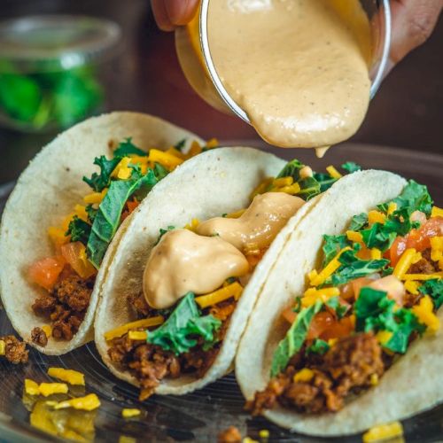 Three tacos filled with ground meat, shredded cheese, and greens, being topped with a creamy sauce.