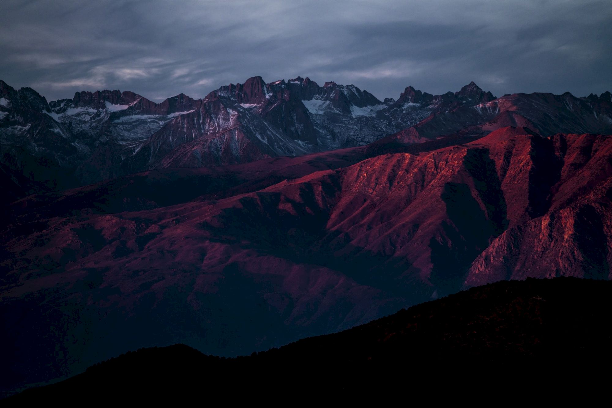 A mountain range is bathed in deep red light under a cloudy sky at dusk or dawn, creating a dramatic and moody landscape.