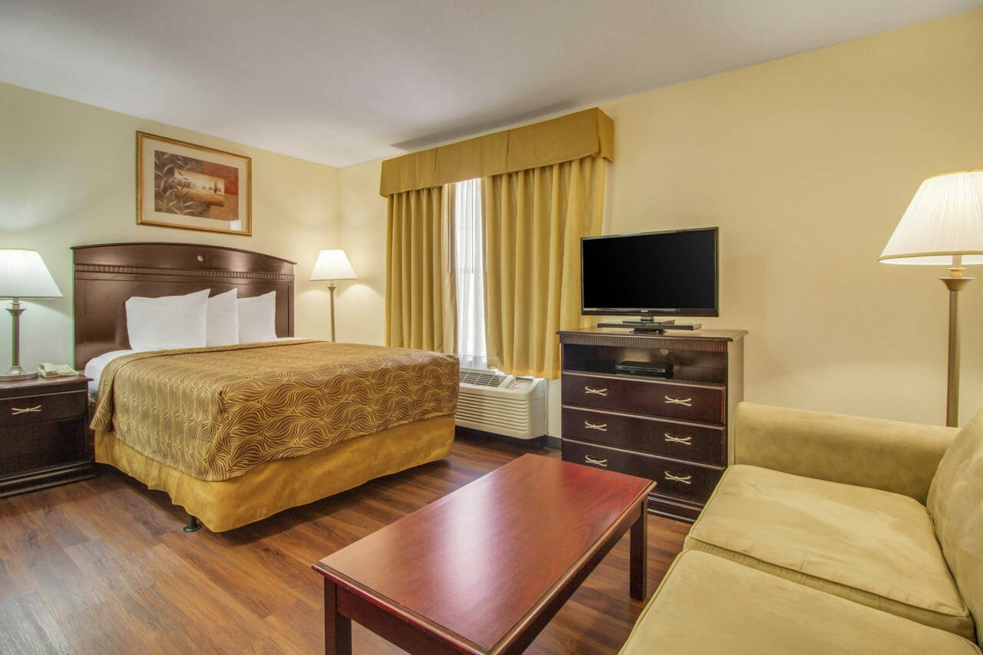 A hotel room with a bed, sofa, TV, lamps, and a wooden floor, decorated in warm tones and simple furnishings.