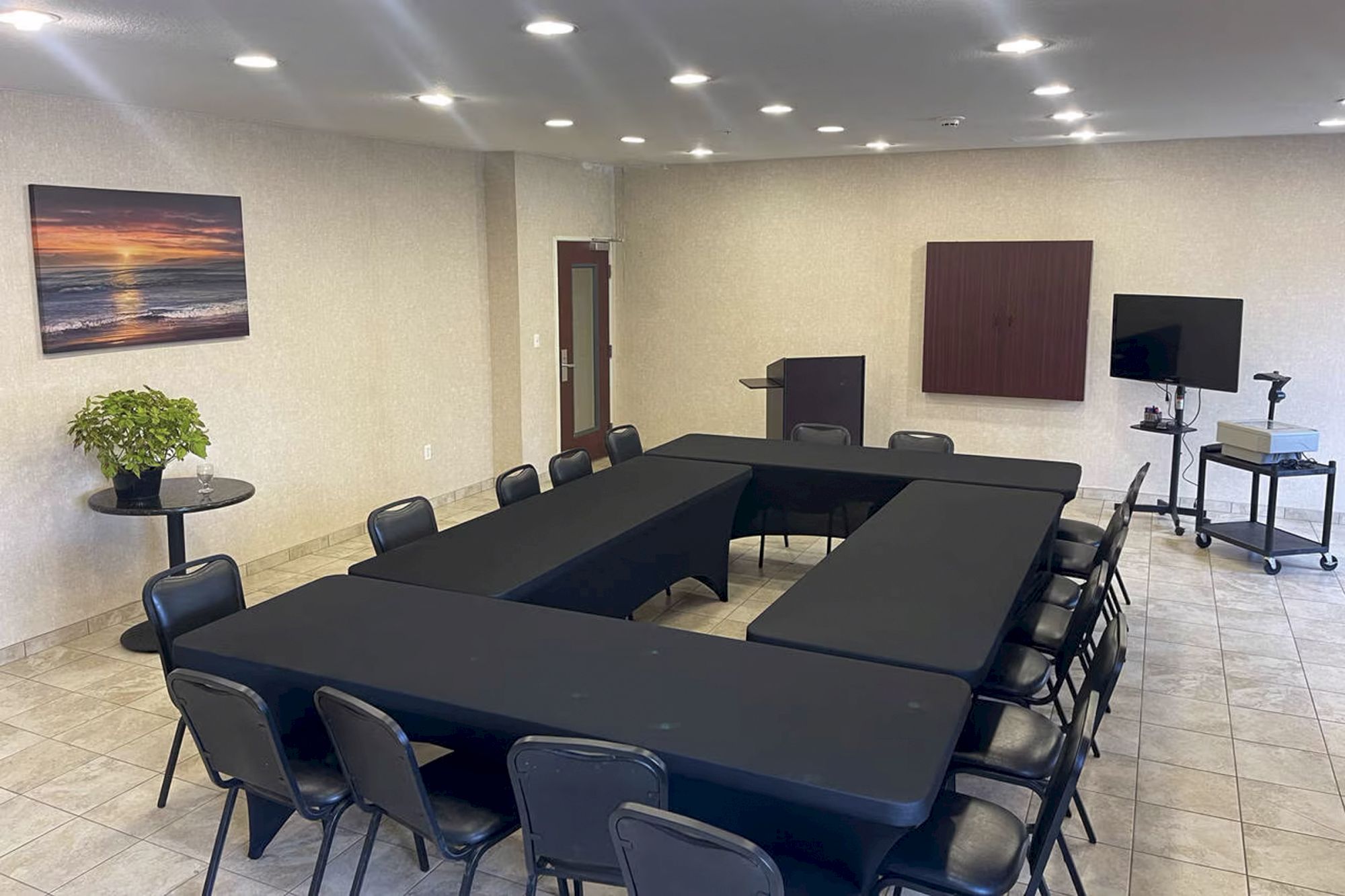 A conference room with black-covered tables arranged in a U-shape, chairs, a podium, TV, and wall art.