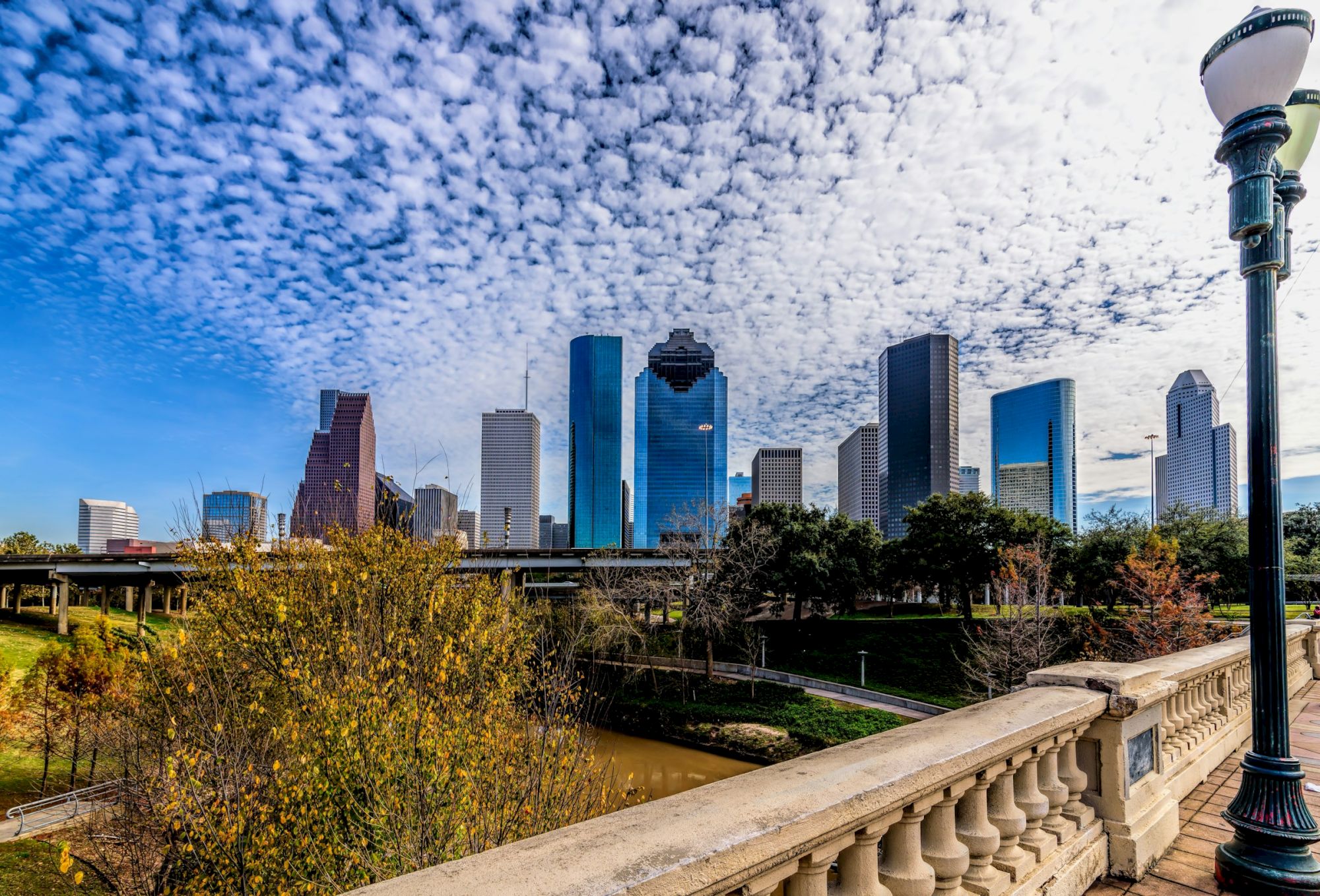 A city skyline with tall buildings under a partly cloudy sky, viewed from a bridge with trees and a river in the foreground.