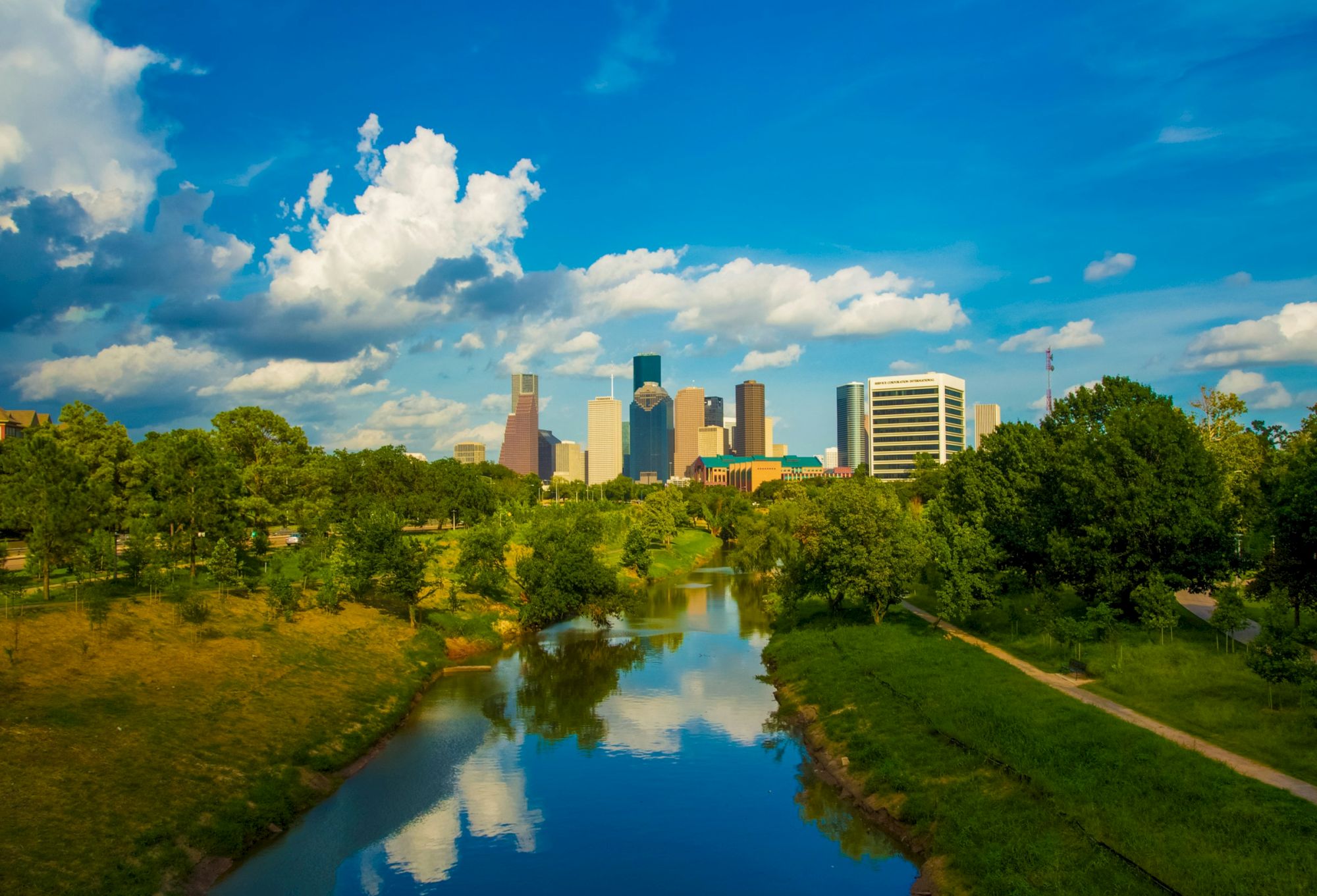 A city skyline with skyscrapers, lush greenery, and a river under a blue sky with clouds is in the image.