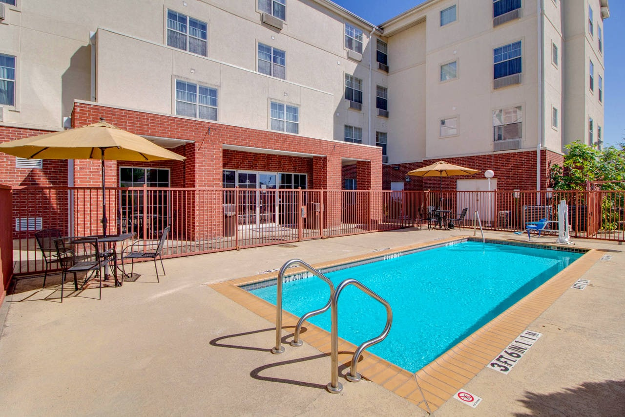 A small outdoor swimming pool with metal ladders, surrounded by a fence, and patio tables with umbrellas near a building.