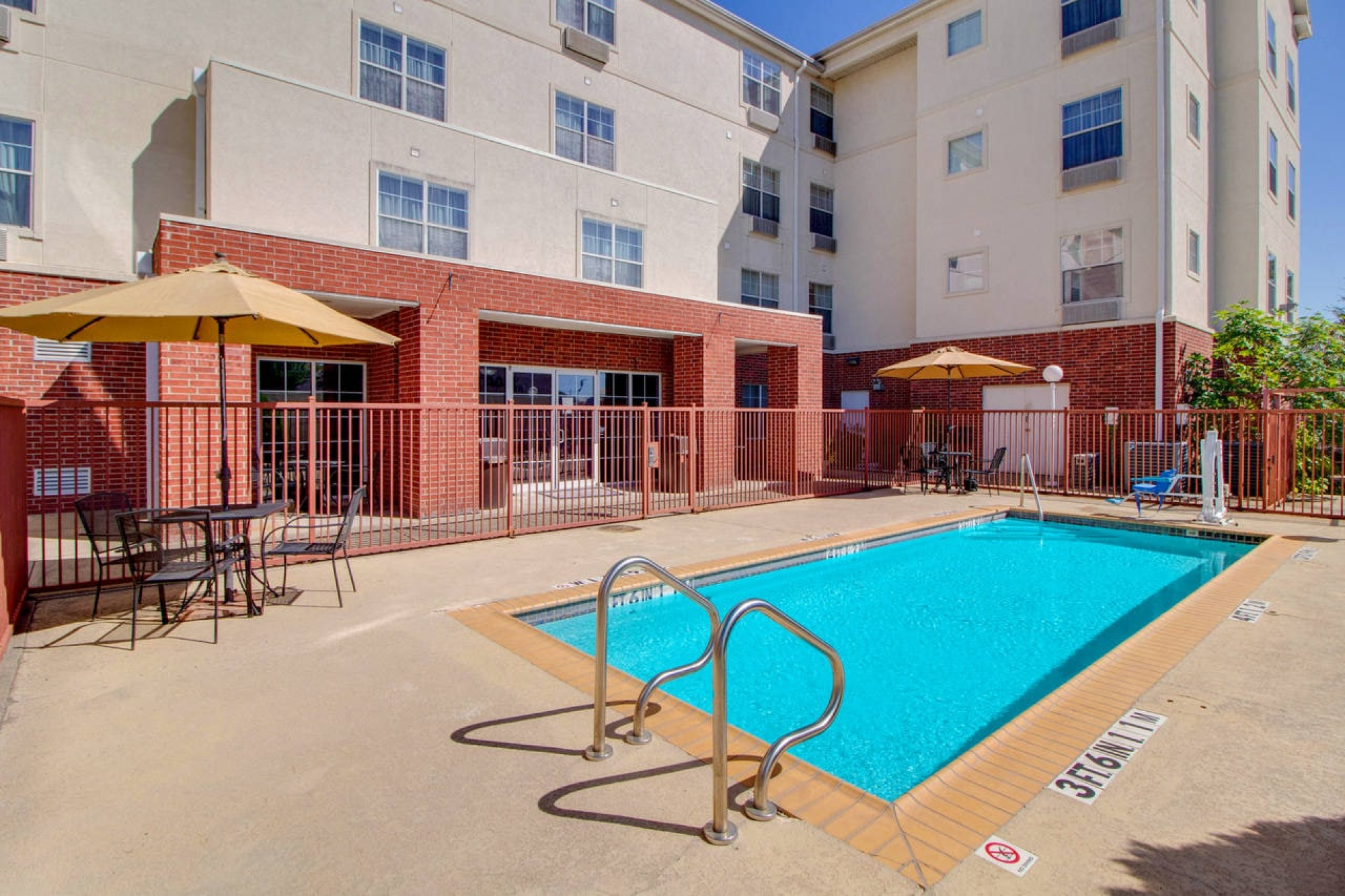 A small outdoor swimming pool with metal ladders, surrounded by a fence, and patio tables with umbrellas near a building.