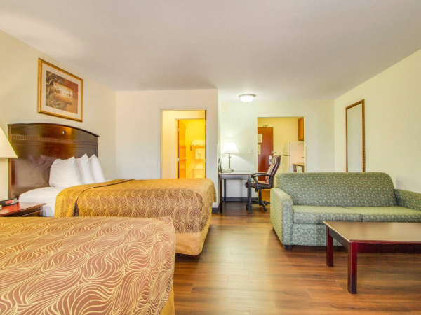 The image shows a hotel room with two beds, a sofa, a table, a desk with a chair, and a lamp, featuring warm colors and wooden flooring.