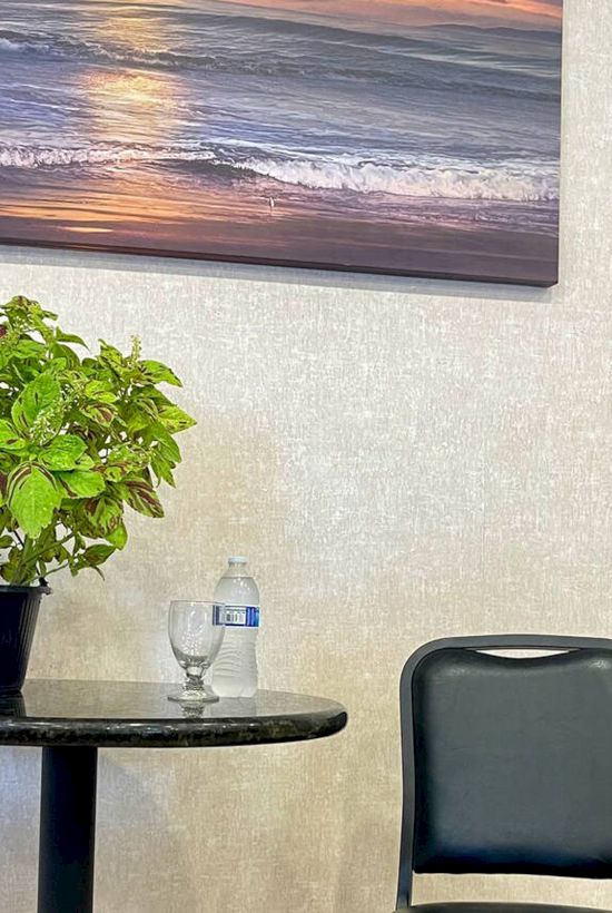 A small table with a potted plant and water bottle, next to a chair. A sunset beach painting hangs on the wall above.