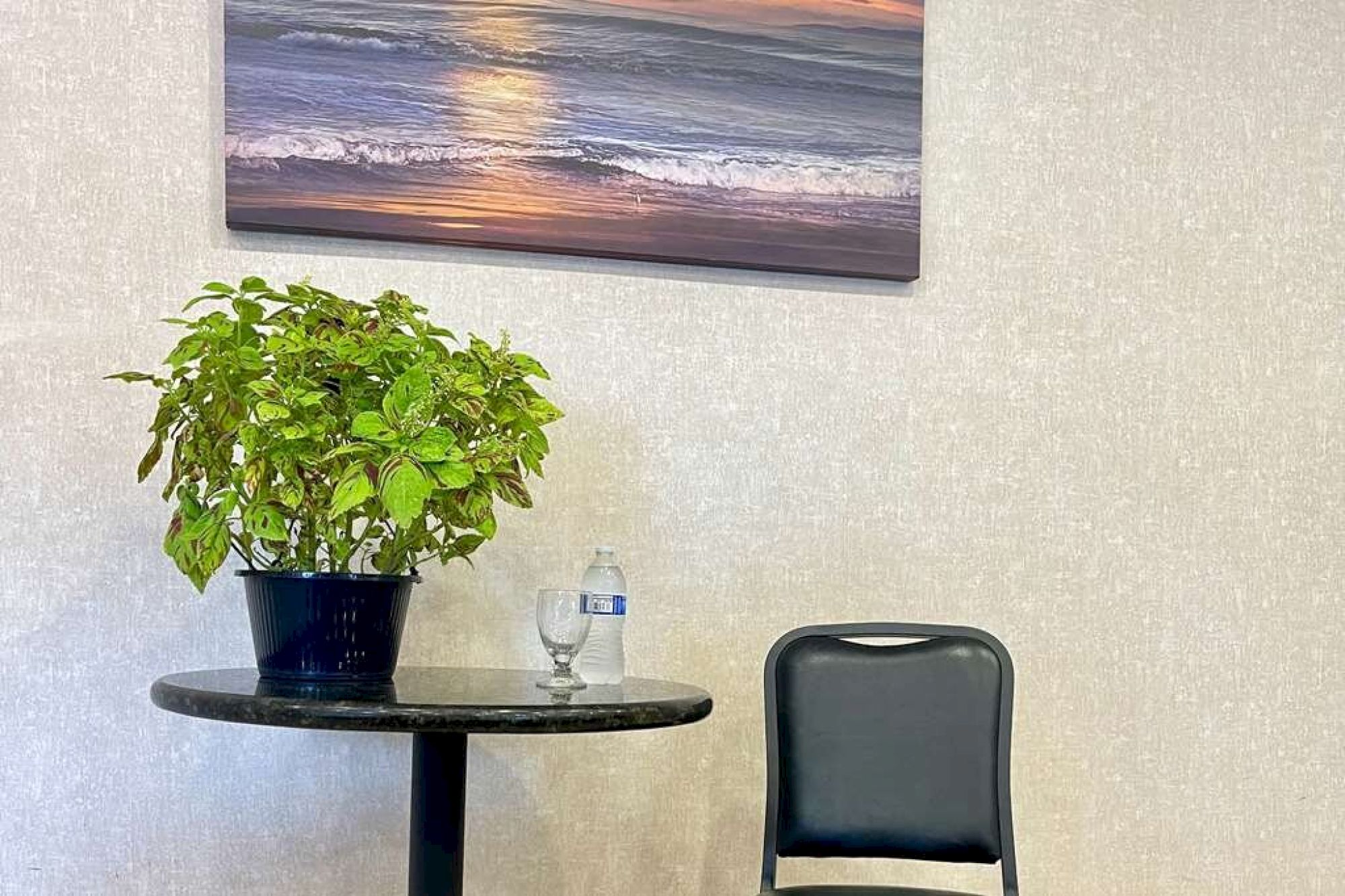 A potted plant and water bottle on a round table, a black chair nearby, and an ocean sunset painting on the wall complete the scene.