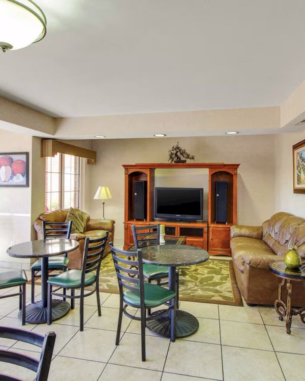 A cozy living area with tables, chairs, a TV, bookshelf, sofas, and decor in a room with large windows and tile flooring.