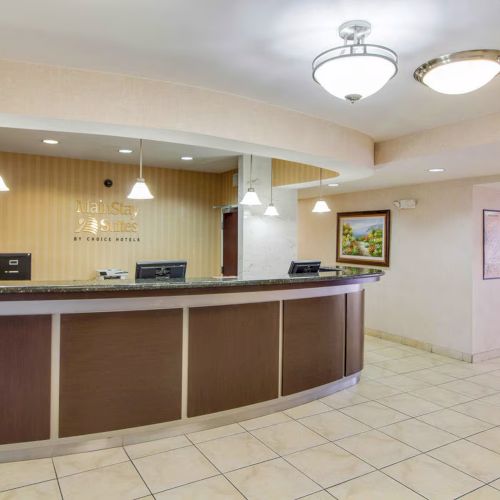 A hotel reception area with a long counter, overhead lights, and a painting on the wall. The floor is tiled, and the area is well-lit.