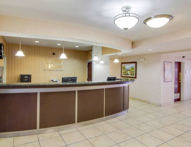 A hotel reception area with a long counter, overhead lights, and a painting on the wall. The floor is tiled, and the area is well-lit.