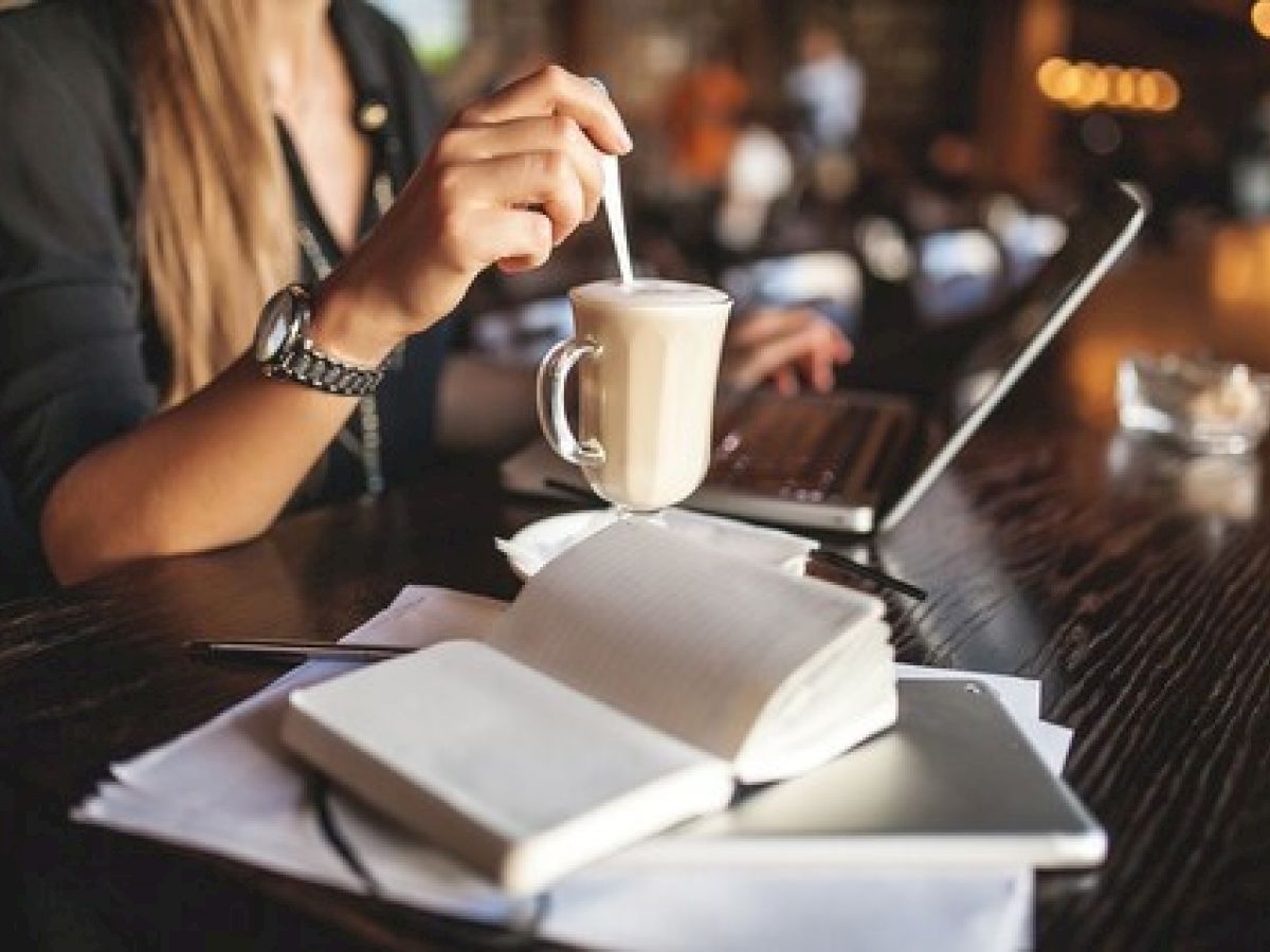 A person in a cafe is stirring a drink while using a laptop, with notebooks and a smartphone on the table.