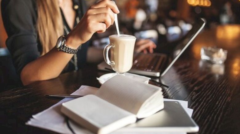 A person in a cafe is stirring a drink while using a laptop, with notebooks and a smartphone on the table.