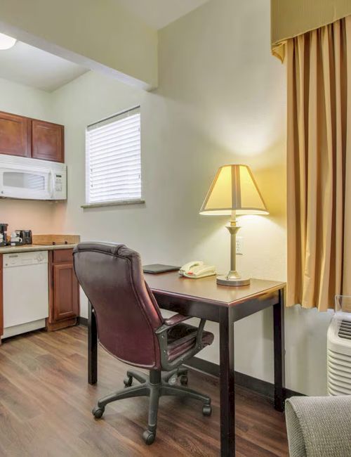 A kitchenette with wooden cabinets, appliances, a lamp on a desk, a chair, and a window with curtains is shown.