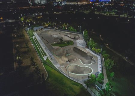 The image shows an illuminated skate park at night with ramps and bowls, surrounded by trees and pathways in a park setting.