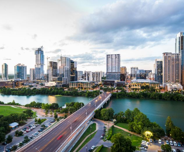 A city skyline with tall buildings, a river, a bridge, and green spaces under a partly cloudy sky.