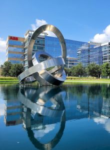 A modern office building with a reflective glass facade and a metallic ring sculpture, set beside a tranquil pond under a clear blue sky.