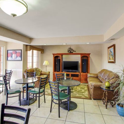 A living area with tables, chairs, and sofas facing a large TV and entertainment center. Natural light comes from sliding doors.