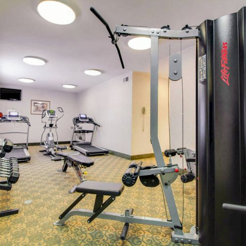 The image shows a small gym with various exercise equipment, including a strength training machine and treadmills.